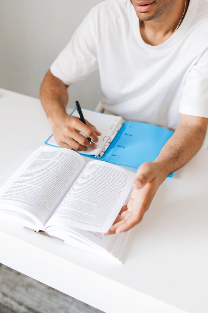 An adult man takes notes from a book while studying at home, writing in a notebook.